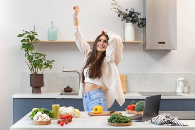 Çekici bir kadın vejetaryen yemeği pişirirken gülümser ve dans eder. Sağlıklı beslenme konsepti, sağlıklı bir yaşam tarzı hakkında blog yazma.