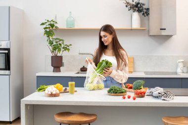 Çekici bir kadın gülümser ve mutfaktaki ağdan taze yiyecek çıkarır. Sıfır atık kavramı, sağlıklı bir yaşam tarzı ve sağlıklı bir diyet..