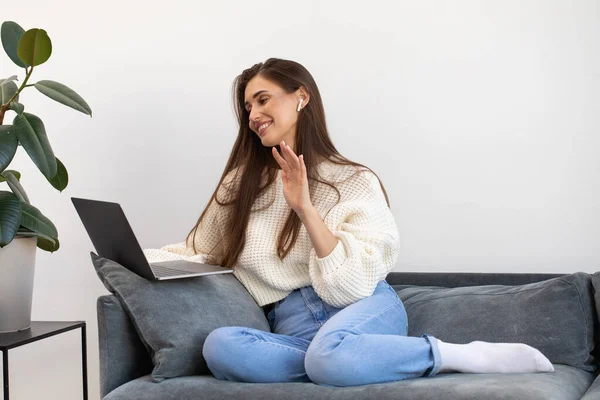 stock image A young woman is sitting on a sofa, smiling and waving during an online meeting using a laptop. The concept of remote work, online learning, meeting at a distance.