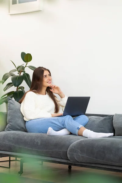 stock image An attractive brunette uses a laptop, sitting on the sofa and smiling dreamily. The concept of online shopping, work at home.