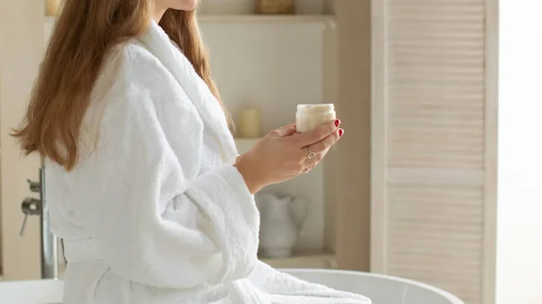 一位穿着白衣的漂亮女人坐在浴室里 手里拿着一罐抗衰老霜 可以使面部和身体的皮肤保湿 日常皮肤护理 — 图库照片