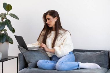 Çekici genç bir kadın, kablosuz kulaklıklarla kanepede rahat bir şekilde oturuyor ve dizüstü bilgisayar kullanıyor. Evde çalışma kavramı, serbest çalışma, online eğitim..