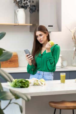 Genç ve çekici bir kadın mutfakta durup yeşil bir elma yerken telefonu kullanıyor. Yaşam tarzı, mutfakta güzel bir esmerin portresi..