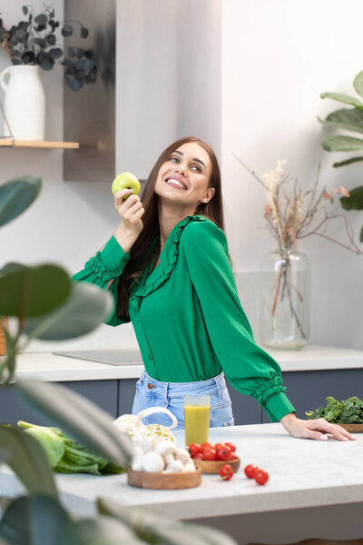 A happy young woman holds a green apple in her hand in the kitchen. The concept of healthy eating, vegetarianism.