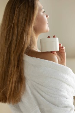 Bir kadın elinde kozmetik, yüz ve vücut kremi olan beyaz bir kavanoz tutuyor ve sırtı kameraya dönük duruyor. Cilt bakımı kavramı.