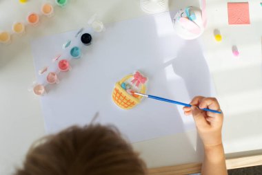 Küçük tatlı bir kız bir masada oturuyor ve Paskalya zanaatı yapıyor, Paskalya heykelciklerini boyuyor. Çocuklar için festival sanatı dersi. Yaratıcılık ve hobiler.