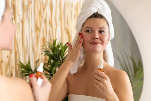 Aynanın önündeki çekici bir kadın banyodaki yüz derisine serum uyguluyor. Cilt bakımı, yaşlanmayı önleyici kozmetik.