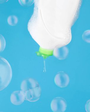 Bir damla bulaşık jeli, uçan sabun köpükleriyle mavi bir zemin üzerinde bir şişeden akar. Bulaşık jeli beyaz plastik şişe. Mutfağı temizleme fikri.
