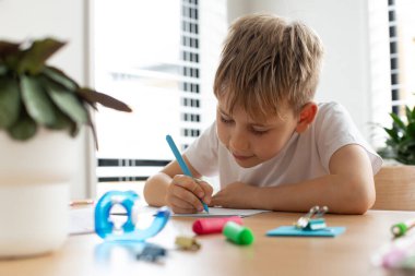 Şirin bir çocuk evdeki masada ödevini yapıyor, bir deftere yazıyor. Evde eğitim, ilkokul. Çocuk eğitimi kavramı.