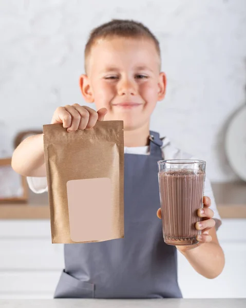 A cute joyful boy holds a glass of cocoa and a craft package with a place for a logo. The concept of cocoa powder and drink.