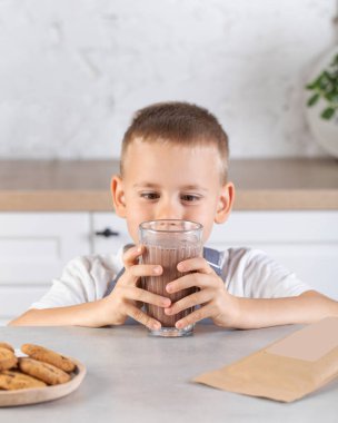 Tatlı bir çocuk mutfaktaki cam bardaktan kakao içiyor. Çikolatalı süt, çocuklar için lezzetli ve sağlıklı bir içecek..