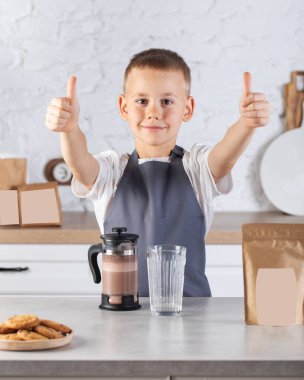 Sevimli bir çocuk mutfak masasında duruyor, pişmiş kakao masanın üstünde ve çocuk başparmağını kaldırıyor. Çikolatalı süt, kakao konsepti.