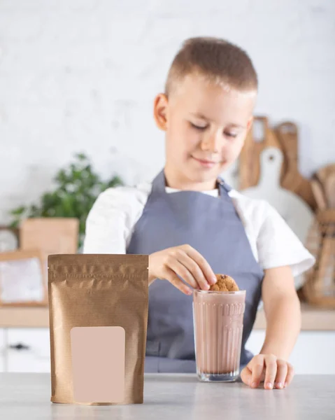 Craft packaging with a place for your logo and a cute boy in the kitchen dips cookies into a glass of cocoa. Advertising of cocoa powder.