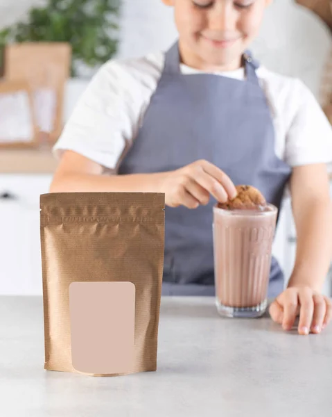 Craft packaging with a place for your logo and a cute boy in the kitchen dips cookies into a glass of cocoa. Advertising of cocoa powder.