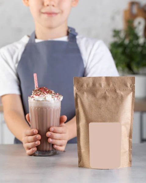 Craft packaging with a place for your logo and a cute boy in the kitchen holding a glass with a chocolate milkshake. Advertising of cocoa powder.