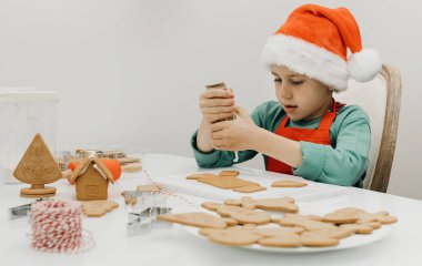 Yakışıklı bir çocuk Noel Baba şapkasıyla Noel Baba 'nın masasında zencefilli kurabiye süslüyor. Çocuklarla Noel tatili.
