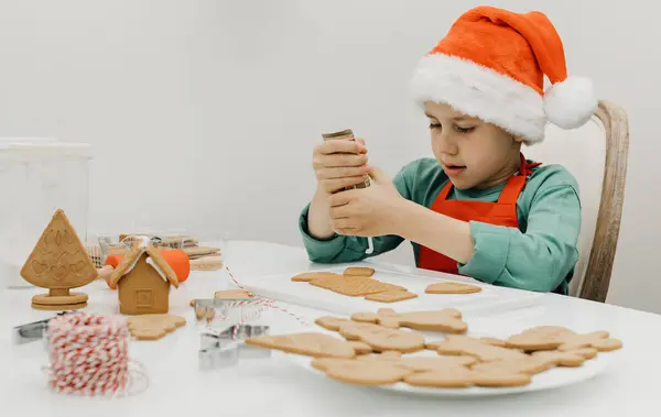 A cute boy decorates gingerbread cookies at the table in Santas hat on New Years Eve. Christmas holidays with children.