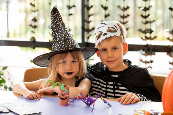 Karnaval kostümlü tatlı bir oğlan ve kız bir masada oturup Cadılar Bayramı kutlamaları için el işi yapıyorlar. Mutlu Cadılar Bayramı.