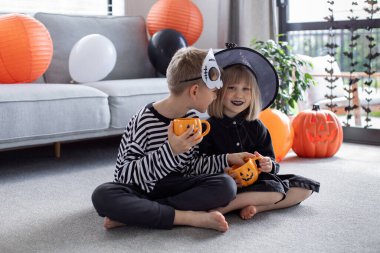 Karnaval kostümlü şirin erkek ve kız kardeşler Cadılar Bayramı partisinde komik bir balkabağı şeklinde fincanlarla yerde oturup eğleniyorlar. Mutlu Cadılar Bayramı.