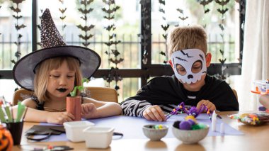 Cadılar Bayramı kostümlü şirin erkek ve kız kardeşler masada şenlikli el sanatları yapıyorlar. Cadılar Bayramı el sanatları üzerine bir master sınıfı. Mutlu Cadılar Bayramı.
