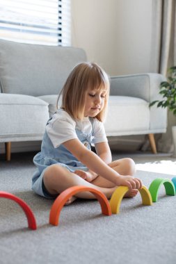 Şirin bir kız ahşap gökkuşağıyla eğitici bir oyun oynuyor, bir odada yerde oturuyor. Çocuklar için ahşap eğitim oyuncağı. Maria Montessoris çocuk gelişimi ve öğrenme metodolojisi.