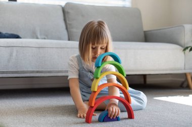 Şirin bir kız ahşap gökkuşağıyla eğitici bir oyun oynuyor, bir odada yerde oturuyor. Çocuklar için ahşap eğitim oyuncağı. Maria Montessoris çocuk gelişimi ve öğrenme metodolojisi.