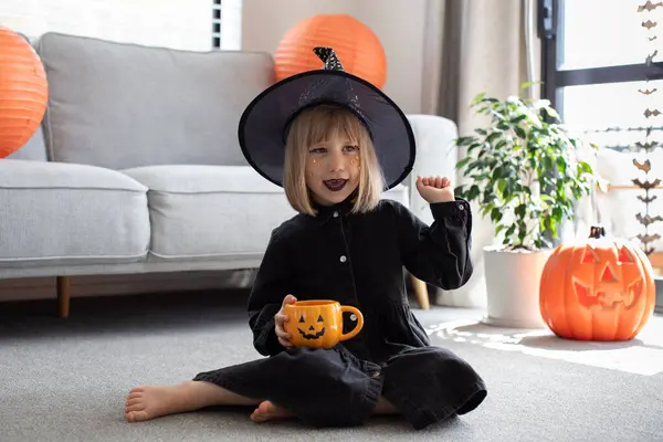 Funny girl in a witch hat at the Halloween party with a pumpkin mug in her hands. The concept of celebrating Halloween with children.