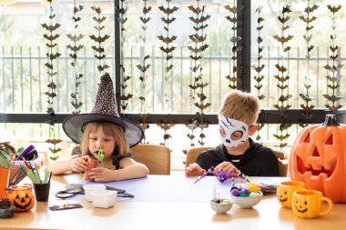 Cadılar Bayramı kostümlü şirin erkek ve kız kardeşler masada şenlikli el sanatları yapıyorlar. Cadılar Bayramı el sanatları üzerine bir master sınıfı. Mutlu Cadılar Bayramı.