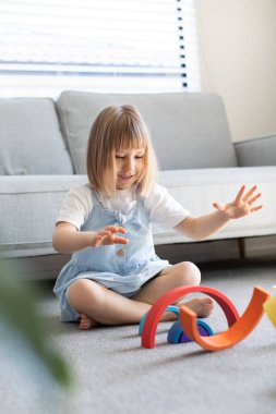 Şirin bir kız ahşap gökkuşağıyla eğitici bir oyun oynuyor, bir odada yerde oturuyor. Çocuklar için ahşap eğitim oyuncağı. Maria Montessoris çocuk gelişimi ve öğrenme metodolojisi.