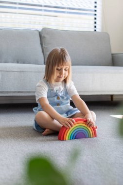 Şirin bir kız ahşap gökkuşağıyla eğitici bir oyun oynuyor, bir odada yerde oturuyor. Çocuklar için ahşap eğitim oyuncağı. Maria Montessoris çocuk gelişimi ve öğrenme metodolojisi.