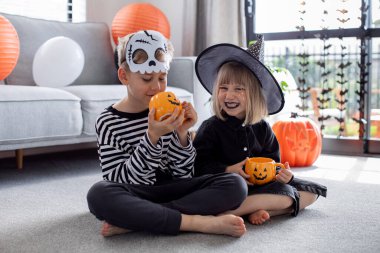 Karnaval kostümlü şirin erkek ve kız kardeşler Cadılar Bayramı partisinde komik bir balkabağı şeklinde fincanlarla yerde oturup eğleniyorlar. Mutlu Cadılar Bayramı.