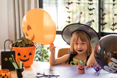 Cadı kostümü giymiş tatlı bir kız Cadılar Bayramı 'nda usta sınıfında elinde balonla masada oturuyor. Cadılar Bayramı partisi.