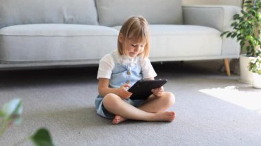 Sevimli bir anaokulu kızı bir odada yerde otururken dijital bir cihaz kullanıyor. Telefon ve tabletler için çocuk eğitim uygulamaları. Çocuklar ve dijital teknolojiler.