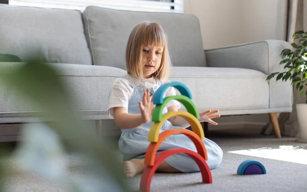 stock image A cute girl is playing an educational game with a wooden rainbow, sitting on the floor in a room. Childrens wooden educational toy. 