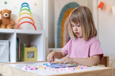 Şirin bir kız çocuk odasındaki bir masada oturuyor ve ponponlardan el işi yapıyor. Çocukların yaratıcılığı, çocuk el sanatları kavramı..