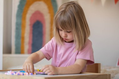Şirin bir kız çocuk odasındaki bir masada oturuyor ve ponponlardan el işi yapıyor. Çocukların yaratıcılığı, çocuk el sanatları kavramı..