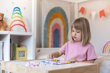 Şirin bir kız çocuk odasındaki bir masada oturuyor ve ponponlardan el işi yapıyor. Çocukların yaratıcılığı, çocuk el sanatları kavramı..