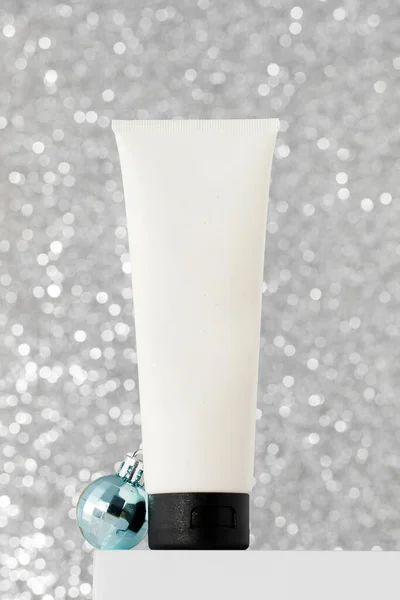 A tube with skin care products on the podium with Christmas balls on a bright silver background. The concept of New Years gifts, protective skin care products in winter.