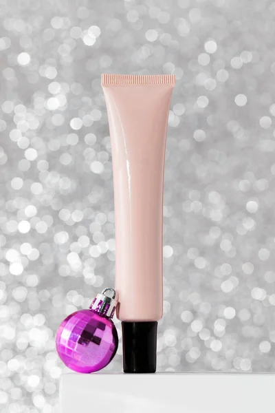 A tube with skin care products on the podium with Christmas balls on a bright silver background. The concept of New Years gifts, protective skin care products in winter.
