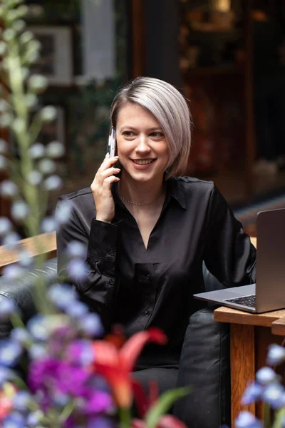 A young business woman working smiles with a phone in her hands, sitting at a laptop in a public place. The concept of a business woman, a freelance woman.