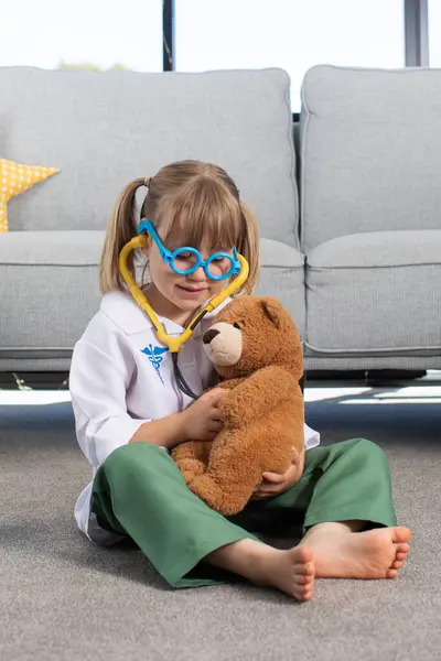 A cute little girl is playing in a doctor costume with a teddy bear. A happy little child is playing at the hospital, at the veterinarian. The concept of healthcare, veterinary medicine.