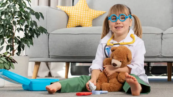 A cute little girl is playing in a doctor costume with a teddy bear. A happy little child is playing at the hospital, at the veterinarian. The concept of healthcare, veterinary medicine.