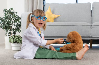 Küçük tatlı bir kız, doktor kostümüyle oyuncak ayıyla oynuyor. Hastanede, veterinerde küçük mutlu bir çocuk oynuyor. Sağlık ve veterinerlik hekimliği kavramı.