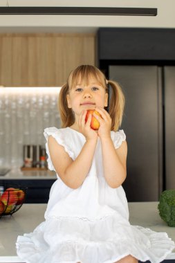 Mutfak masasında elinde kırmızı bir elmayla sevimli bir kız oturuyor. Sağlıklı beslenme kavramı.