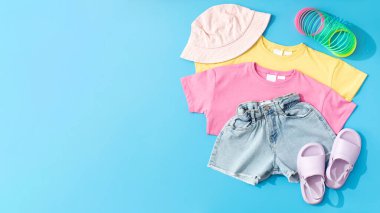 Yazlık moda kıyafetlerinden oluşan bir çocuk seti, mavi arka planda deniz gezisi için bir görüntü. Düz yatıyordu. Denim şortlar, renkli tişörtler ve kızlar için parlak aksesuarlar..