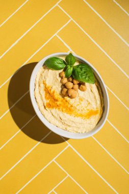 Sarı fayans masasında servis edilen nohut ve fesleğenle kaplı bir humus tabağı.