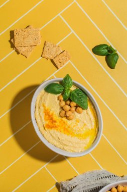 Sarı fayans masasında servis edilen nohut ve fesleğenle kaplı bir humus tabağı.