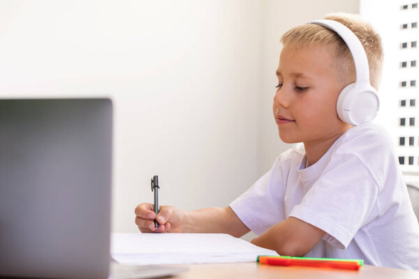 A cute boy is studying at an online school. The boy is sitting in front of a laptop with headphones and listening to the teacher. The concept of education.