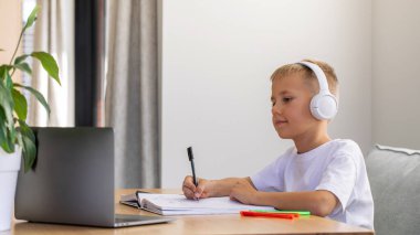 Sevimli bir çocuk çevrimiçi bir okulda okuyor. Çocuk bir dizüstü bilgisayarın önünde kulaklıkla oturuyor ve öğretmeni dinliyor. Eğitim kavramı.
