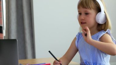 Neşeli küçük kız el sallıyor ve evde online derste resim çizerek konuşuyor. Küçük öğrenci görevlerini yapar ve öğretmenlerin sorularını dizüstü bilgisayarla cevaplar.